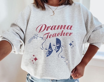 May include: Gray sweatshirt with the text "Drama Teacher" in red and blue lettering. The sweatshirt features two stylized masks in blue and white.