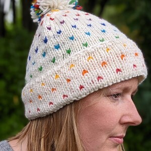 May include: A cream-coloured knitted beanie hat with a rainbow chevron pattern. The hat has a folded brim and a fluffy pom-pom with rainbow detailing. This warm hat is designed for style and comfort.