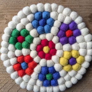 May include: A round, colorful trivet made of felted wool balls. The trivet features a white border with vibrant flower-like designs in red, yellow, green, blue, and purple. The trivet is designed to protect surfaces from heat.