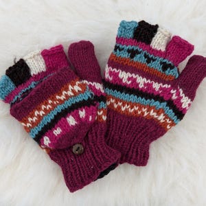 Puede incluir: Un par de guantes sin dedos de punto en tonos burdeos, rosa, azul y blanco. Los guantes presentan un diseño estampado y un cierre de botón en la solapa de la manopla. Se muestran sobre una superficie blanca y esponjosa.