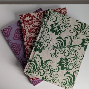 May include: Three notebooks with different patterned covers. The top notebook has a green and white floral design, the middle notebook has a red and white floral design, and the bottom notebook has a purple and white floral design.