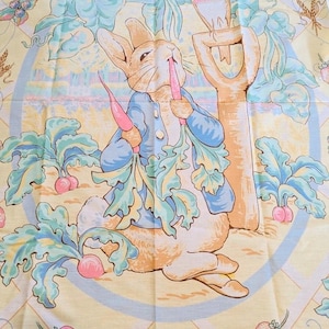May include: A Peter Rabbit themed fabric with a light yellow background. The illustration features Peter Rabbit in a blue jacket, holding carrots, with a bird perched on a shovel. The fabric is decorated with strawberries and vegetables.