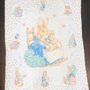 May include: A white quilted blanket featuring illustrations of Peter Rabbit and other characters from Beatrix Potter's stories. The blanket has a floral pattern and a blue border with the text "Beatrix Potter" at the bottom.