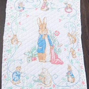 May include: White quilted fabric featuring illustrations of Peter Rabbit and other characters from Beatrix Potter's stories. The design includes a central image of Peter Rabbit in a blue jacket, surrounded by floral and vine motifs.