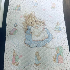 May include: A quilted baby blanket featuring illustrations of Beatrix Potter characters. The central image shows a mouse in a blue dress with two babies. Other characters include rabbits and a duck, each holding a sign. The blanket is white with a floral pattern.