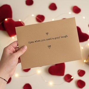 Puede incluir: Una tarjeta de papel kraft con el texto "Open when you need a good laugh" y símbolos de corazón. La tarjeta está rodeada de decoraciones rojas en forma de corazón, pétalos de rosa y una pequeña caja de regalo con una cinta roja.