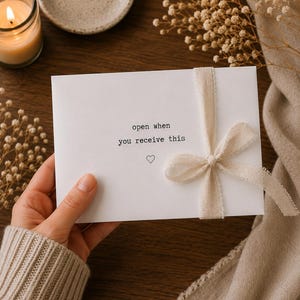 May include: A white rectangular envelope featuring the black printed text open when you receive this and a small heart icon. The envelope is tied with a light beige frayed ribbon tied into a bow on the right side of the front surface.