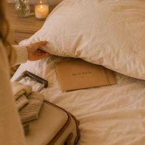 May include: A rectangular tan paper envelope with a string closure, featuring the black printed text Open On Day One and a small heart symbol, resting on a beige bed sheet beneath a large pillow next to a suitcase and folded knit clothing.