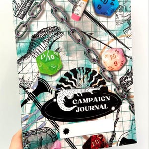 May include: A black and white illustration of a dragon with the text "Campaign Journal" on a black background. The journal cover has a grid pattern with a chain, a pencil, and several dice in different colors.