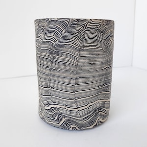 May include: A cylindrical ceramic vase with a black and white striped pattern. The pattern resembles wood grain.