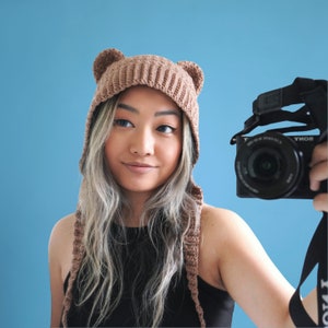 Może przedstawiać: Kobieta w brązowej, dzianej czapce z uszami w kształcie niedźwiedzia, z długimi klapami na uszy, patrząca w obiektyw aparatu, trzymając czarny aparat.