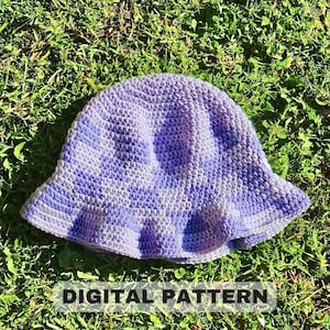 May include: A purple and white crocheted bucket hat with a wide brim, lying on a bed of green grass. The text "DIGITAL PATTERN" is printed below the hat.