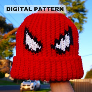 May include: Red crochet beanie with black and white eyes, resembling a superhero character. The beanie has a ribbed texture and a folded brim.