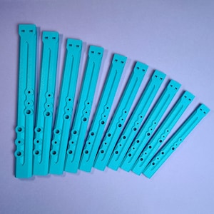 以下が含まれることがあります： ターコイズ色の長方形の楽器部品、おそらくフルートまたはリコーダーが10個、扇状に配置されています。各部品には一連の丸い穴と上部に長方形の開口部があります。オブジェクトは固体材料でできており、均等に配置されています。