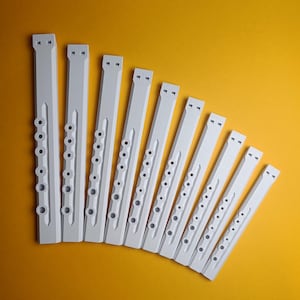 以下が含まれることがあります： 黄色い背景に斜めに配置された、10個の白い長方形の楽器部品。おそらくフルートまたはリコーダー。各ピースには一連の丸い穴と上部に長方形の開口部があります。