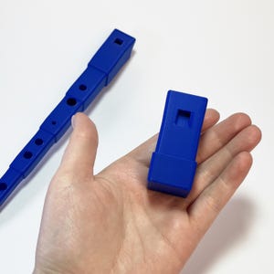 Può includere: Un flauto di plastica blu con sei fori e un bocchino. Il flauto è smontato e i pezzi sono disposti su una superficie bianca.