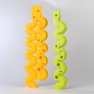 May include: Two abstract sculptures. One is yellow, the other is lime green. Both have a wavy, serpentine design with rounded edges and small circular cutouts. The yellow sculpture has orange accents. The background is plain white.