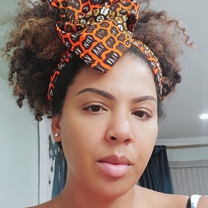 Puede incluir: Una mujer con el pelo rizado castaño lleva una cinta para la cabeza con estampado naranja y marrón brillante. La cinta está anudada en la parte superior de la cabeza. Lleva un collar plateado con pequeñas piedras redondas.