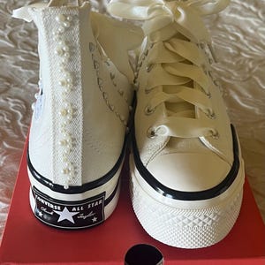 May include: White high-top Converse sneakers with pearl embellishments and a white satin ribbon tied around the laces. The shoes are on a red box with the Converse logo.