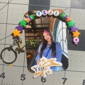 May include: A keychain with a photo card featuring a woman with pink hair. The card has a gold star with the name "YEJI" on it. The keychain has a beaded bracelet with colorful stars and the letters "YEJI" on it.