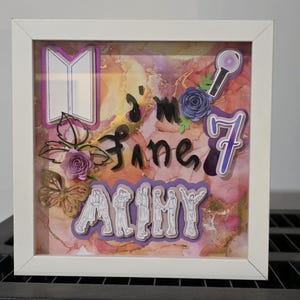 May include: A white framed shadow box with a colorful abstract background. The box features paper cutouts of a book, a microphone, the number 7, and the word "ARMY". The phrase "I'm fine" is written in cursive. Decorative paper flowers and a butterfly are also included.