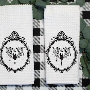 May include: Two white kitchen towels with a black and white design of a bat in a decorative frame. The towels are on a black and white checkered background.