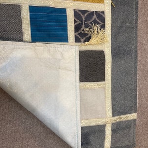 May include: A gray and white patchwork rug with a fringe border. The rug is made of different fabrics and has a variety of patterns. The rug is folded in half and the fringe is visible on the bottom edge.