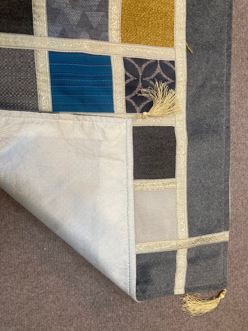May include: A gray and white patchwork rug with a fringe border. The rug is made of different fabrics and has a variety of patterns. The rug is folded in half and the fringe is visible on the bottom edge.