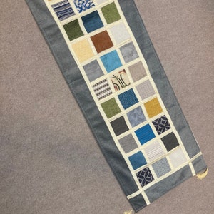 May include: A gray table runner with a patchwork design of various colored squares. The squares are made of different fabrics and patterns, including stripes, floral prints, and geometric designs.