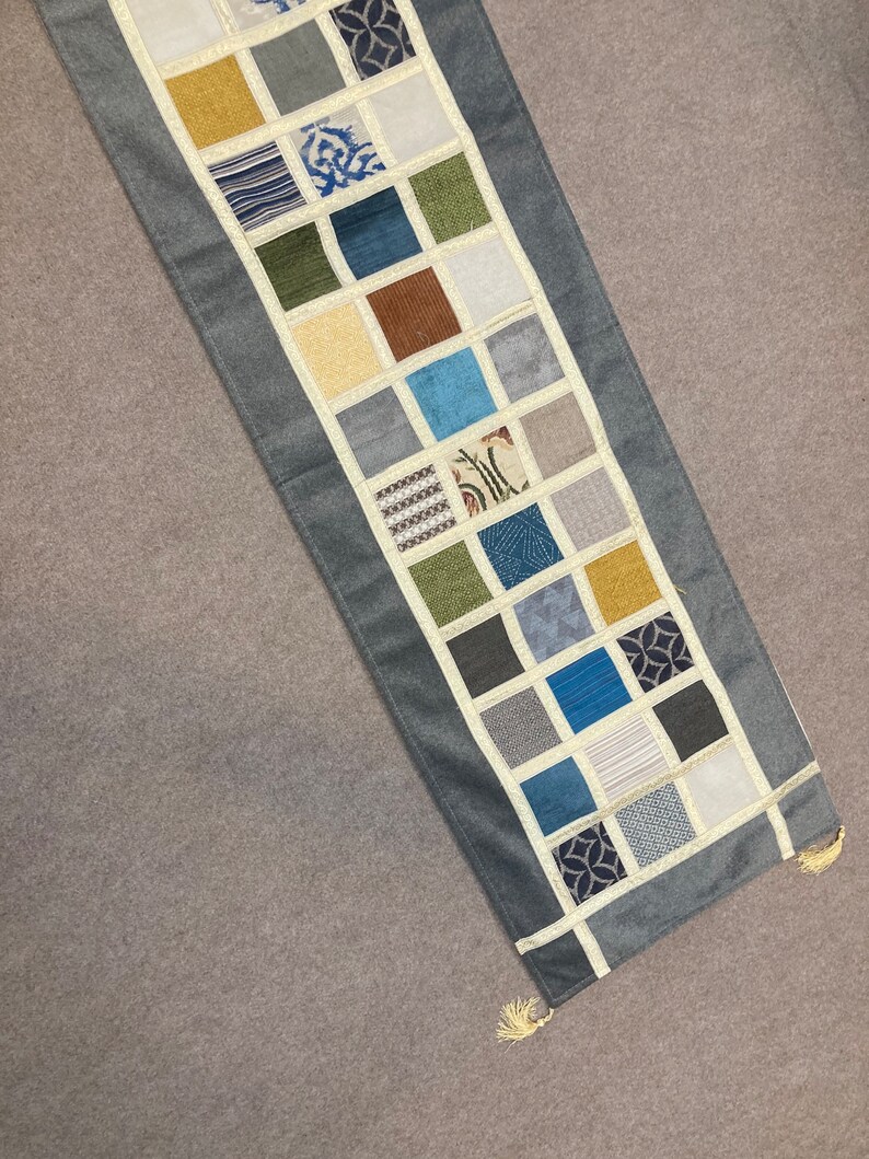 May include: A gray table runner with a patchwork design of various colored squares. The squares are made of different fabrics and patterns, including stripes, floral prints, and geometric designs.