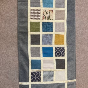 May include: A gray table runner with a patchwork design featuring various patterns and colors, including brown, blue, green, and yellow. The runner has a fringe trim on each end.