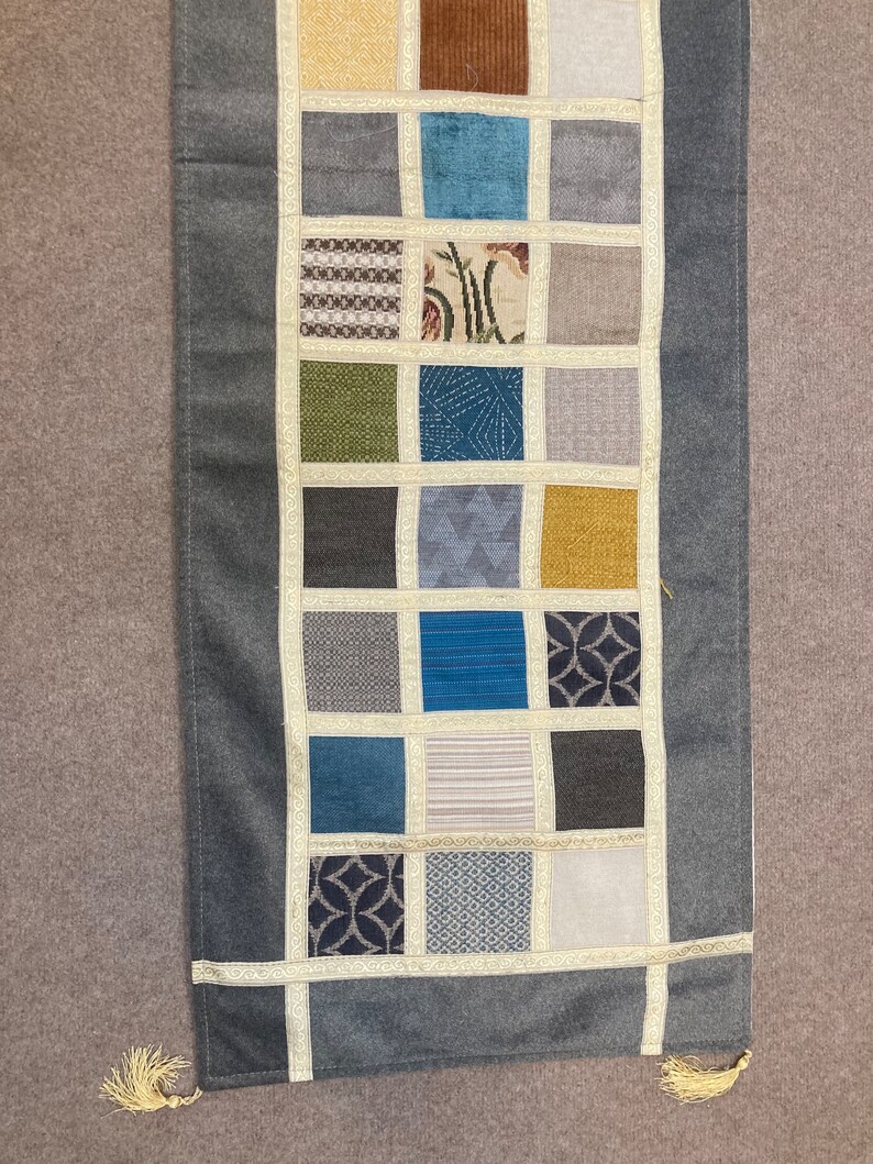 May include: A gray table runner with a patchwork design featuring various patterns and colors, including brown, blue, green, and yellow. The runner has a fringe trim on each end.