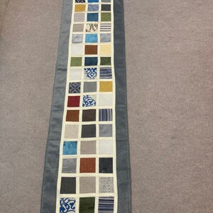 May include: A long, gray table runner with a white border and a patchwork design of 25 squares. Each square is a different fabric with various colors and patterns, including blue, green, brown, and white.