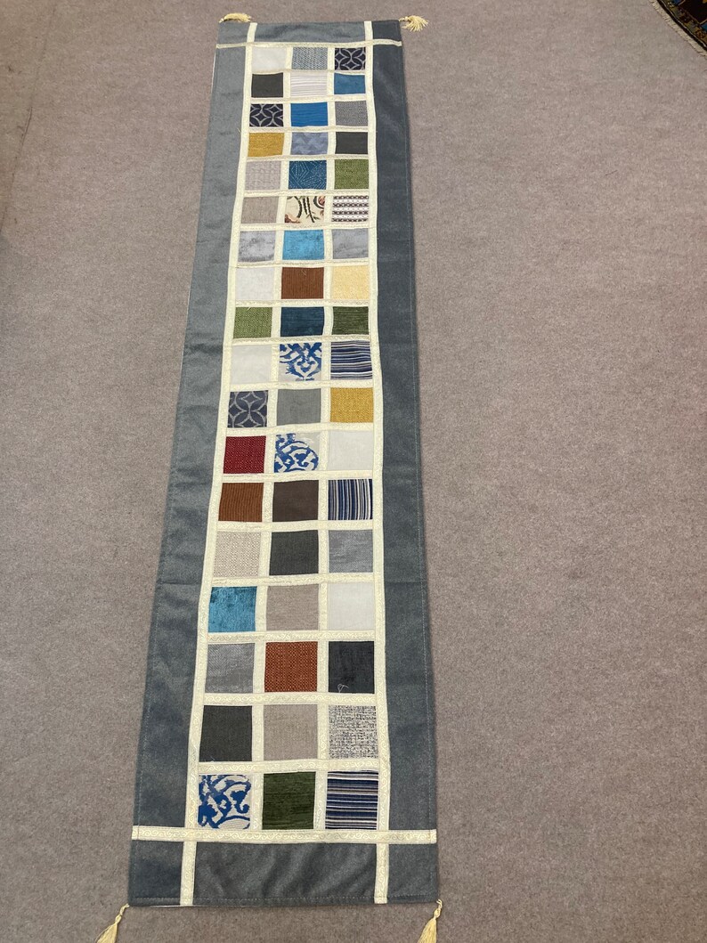 May include: A long, gray table runner with a white border and a patchwork design of 25 squares. Each square is a different fabric with various colors and patterns, including blue, green, brown, and white.