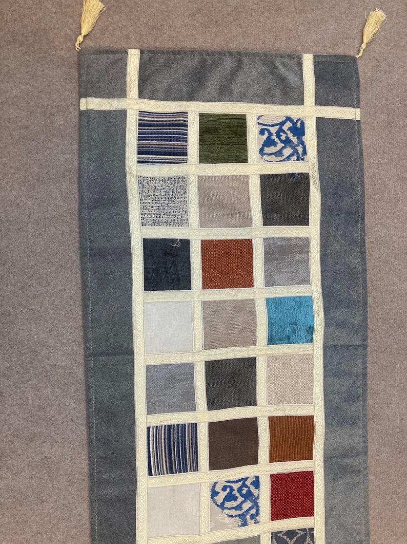 May include: A gray rectangular runner rug with a patchwork design. The rug is made of different fabrics in various shades of gray, brown, blue, and white. The rug has a fringe on each end.