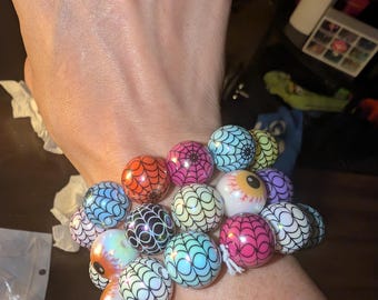 May include: A multi-strand bracelet composed of round beads featuring various colors including orange, pink, blue, and green, each decorated with a black spiderweb pattern, interspersed with white beads featuring a realistic human eye graphic.