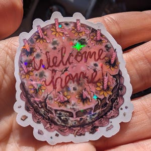 May include: A sticker featuring a round cake design with pink frosting and the words "welcome home" in cursive. The cake is decorated with flowers and candles. The sticker has a holographic effect.