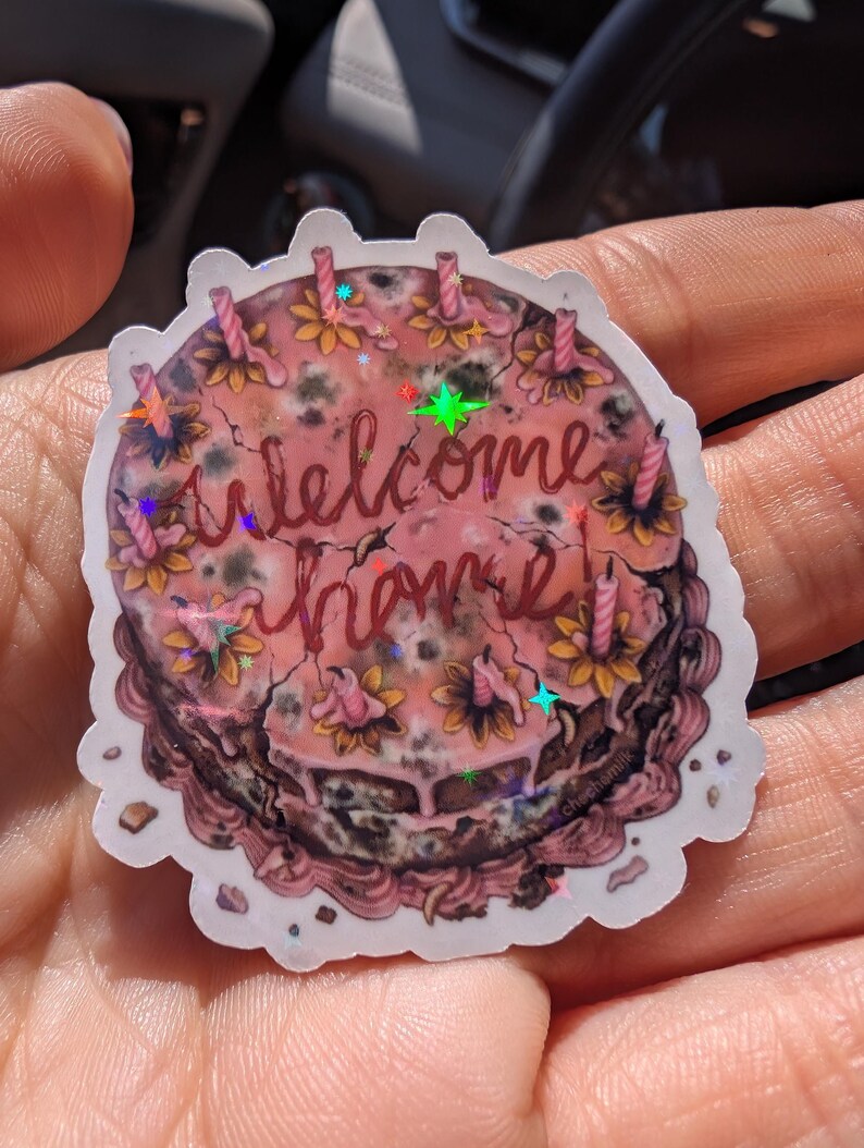 May include: A sticker featuring a round cake design with pink frosting and the words "welcome home" in cursive. The cake is decorated with flowers and candles. The sticker has a holographic effect.