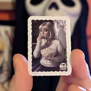 Puede incluir: Una pegatina en blanco y negro que representa a una mujer comiendo palomitas de maíz. La pegatina tiene un borde blanco y un pequeño logotipo. Un máscara de Scream es visible en el fondo. La pegatina es sostenida por una persona.