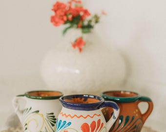 May include: Three handcrafted ceramic mugs with colorful floral designs. The mugs feature white, brown, and blue bases with vibrant accents of red, green, and blue. A white vase with red flowers is in the background.