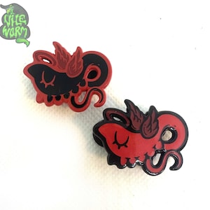 May include: Two red and black enamel pins shaped like a cartoon octopus with wings. The octopus has closed eyes and a small mouth.