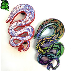 May include: Two enamel pins shaped like a snake with a detailed design. The snake on the left is red with white and blue details. The snake on the right is black with green, yellow, and purple details.
