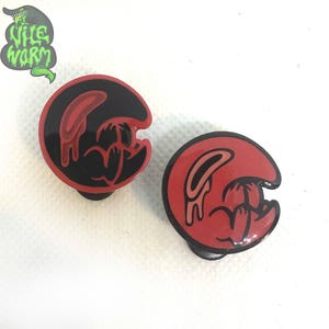 May include: Two enamel pins, one black and one red, with a dripping, cartoon-like figure in the center. The figure is in a half-circle shape and has a black outline.