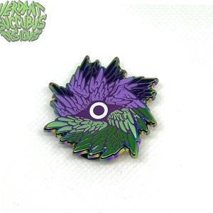 May include: A rainbow iridescent enamel pin featuring a stylised eye with wings. The eye is white with a purple circle in the centre. The wings are purple, green and black.