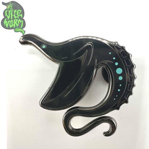 Puede incluir: Pin de esmalte negro con forma de serpiente marina con detalles turquesa. El pin tiene un acabado metálico brillante y el texto "Wile Worm" en un bocadillo verde.