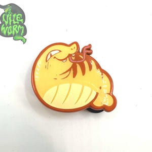 May include: A cartoon-style pin featuring a yellow and brown creature with a tongue sticking out. The pin has a rounded shape with a brown border. The text "The Nile Worm" is above the pin in green.