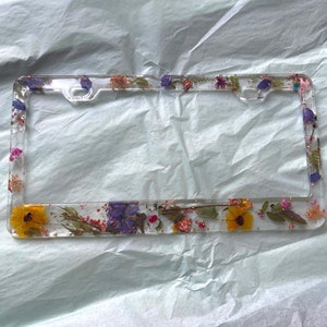 May include: A clear plastic license plate frame with a pressed flower design. The frame is decorated with purple, yellow, and pink flowers, as well as green leaves and gold glitter.
