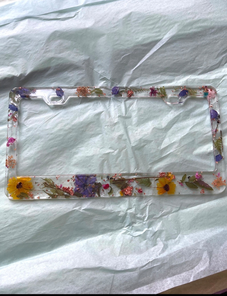 May include: A clear plastic license plate frame with a pressed flower design. The frame is decorated with purple, yellow, and pink flowers, as well as green leaves and gold glitter.
