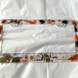 May include: A clear plastic license plate frame with a pressed flower design. The frame features dried flowers, leaves, and gold accents.