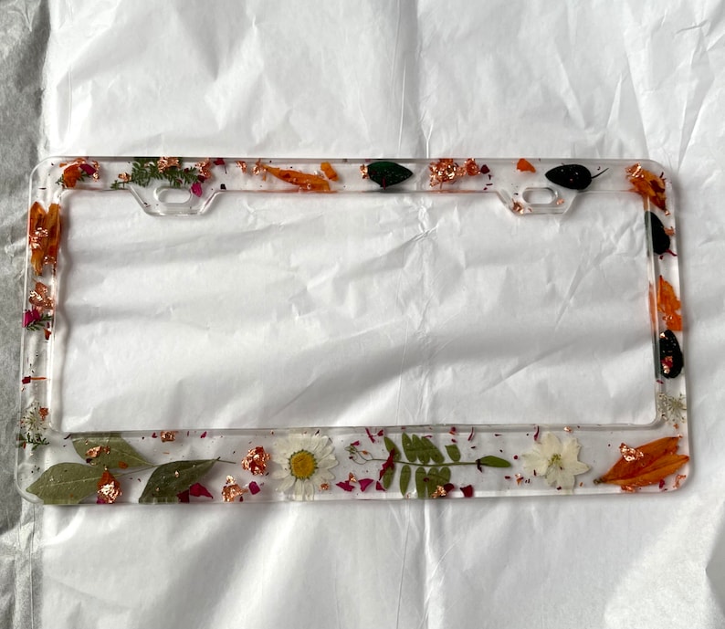 May include: A clear plastic license plate frame with a pressed flower design. The frame features dried flowers, leaves, and gold accents.